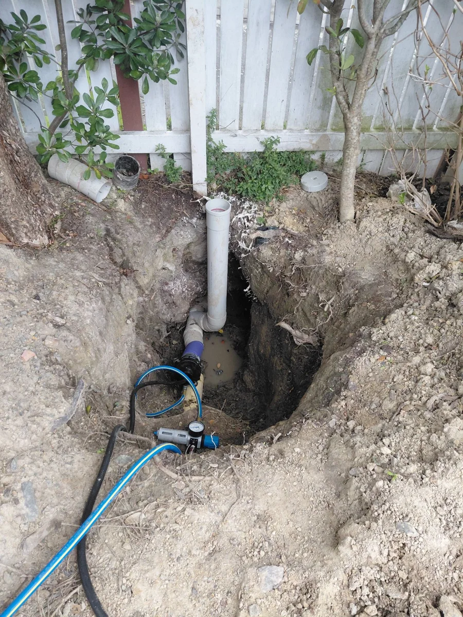Excavated access area with exposed pipework and hoses beside a timber fence in Khandallah